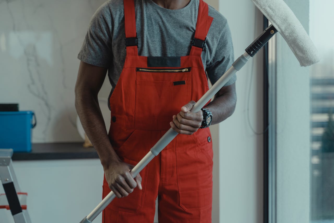 hero-img-02 A professional cleaner wearing red overalls is holding a mop while cleaning indoors.